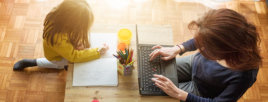 Mother and daughter sit together one playing with toys and one working on a laptop
